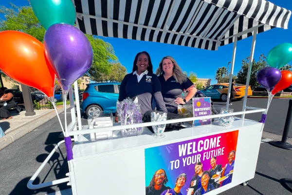 Two smiling women standing behind a white event cart decorated with a black-and-white striped awning and colorful balloons, featuring a sign that reads "Welcome to Your Future."