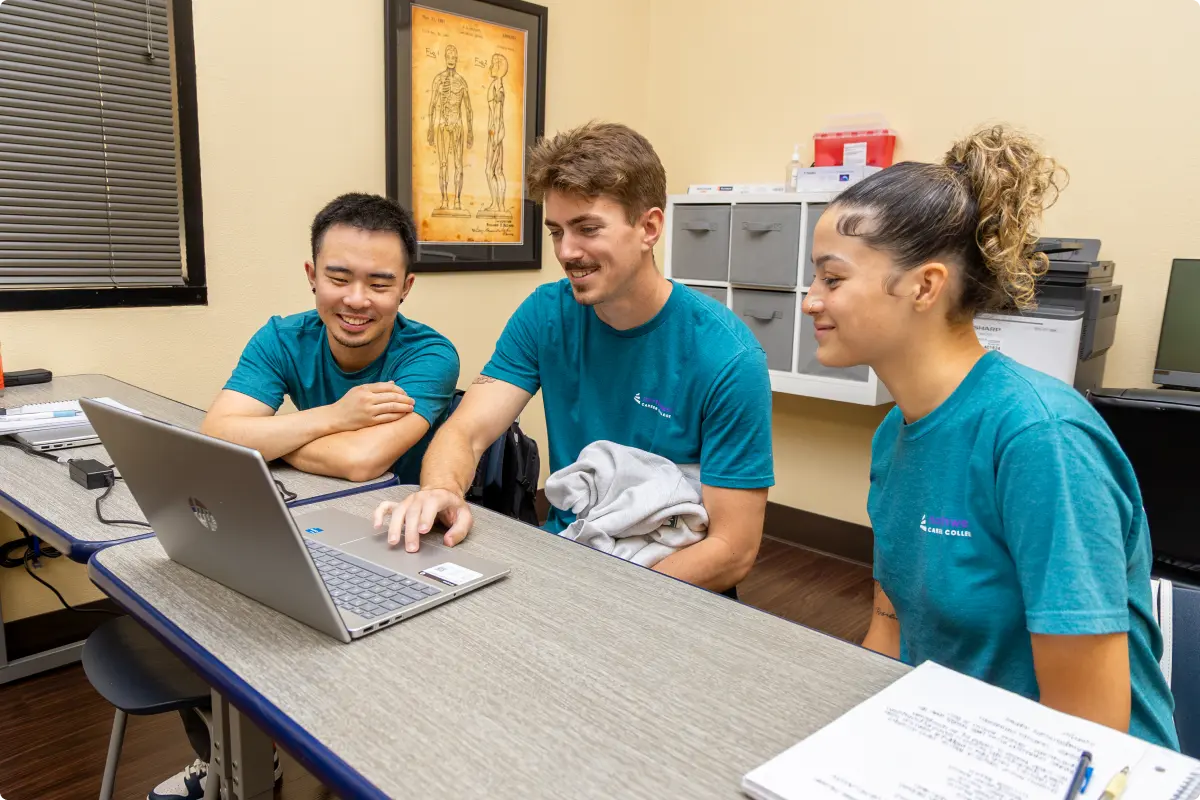 Three Northwest Career College students wearing matching teal shirts smile while looking at a laptop together during the first day of the Summer-4C260706 July class session in a classroom setting.