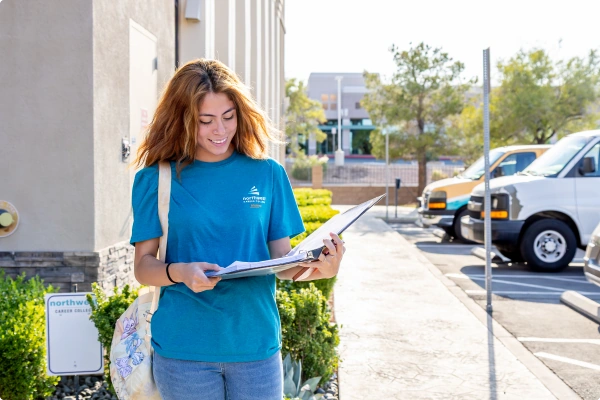 A young woman in a blue shirt smiles while reading a map outdoors, with vehicles and greenery in the background.