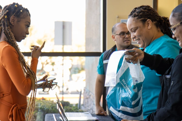 A woman in an orange dress interacts with staff at a table, while another staff member holds a bag and documents.