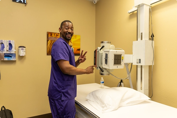 A smiling medical professional in purple scrubs stands by an X-ray machine, giving a peace sign in a clinical room.