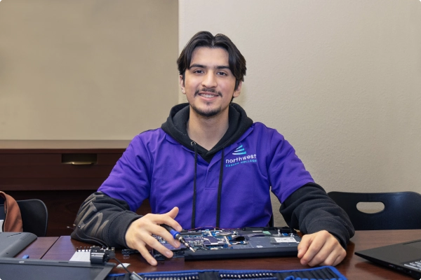 A young man in a purple hoodie smiles while working on a laptop motherboard at a table.