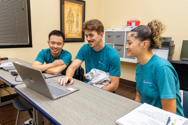 Three students in teal shirts gather around a laptop, engaged in discussion in a classroom setting.