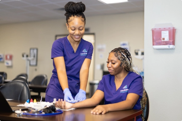 Two women in scrubs are performing a medical procedure in a clinic setting, with equipment on the table.