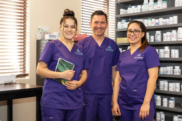 Three healthcare professionals in purple scrubs smile together in a pharmacy setting with shelves of medication behind them.