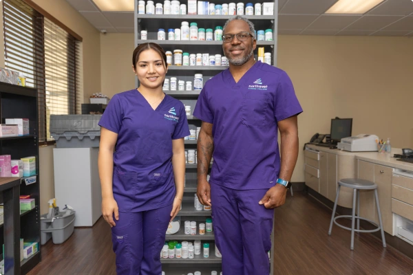 Two healthcare professionals in purple scrubs stand in front of a shelf filled with medication bottles in a pharmacy.