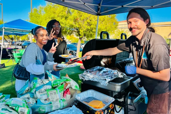 Three people at a food stall, one serving food from a grill, with bags and containers of food in the foreground.
